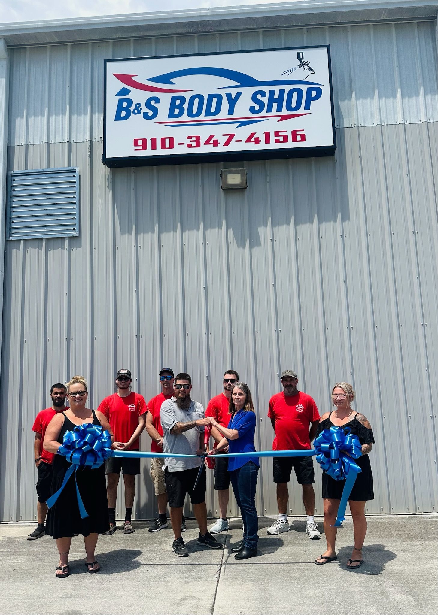 People at a ribbon-cutting ceremony in front of B&S Body Shop with a blue and white sign, and a telephone number.
