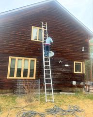 A person stands on a tall extension ladder against the side of a dark wooden house to work on the exterior wall.