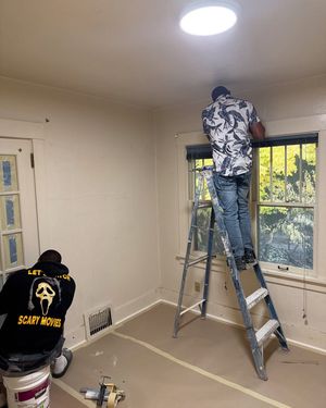 Two people paint the interior of a room, one standing on a ladder by a window and one working on a wall near the floor.