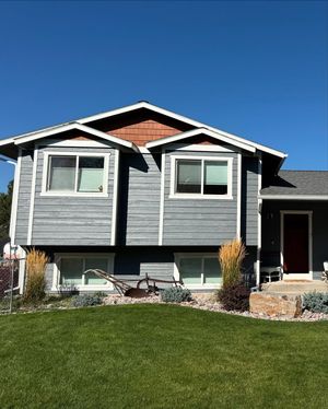 A gray split-level house under a clear blue sky, featuring white trim, two upper windows, and a red front door.