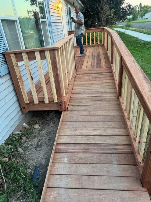 A man stands on a newly constructed wooden wheelchair ramp with railings attached to the side of a house.