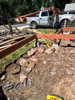 A worker carries a wooden beam near a partially constructed deck in a yard with a white pickup truck in the background.