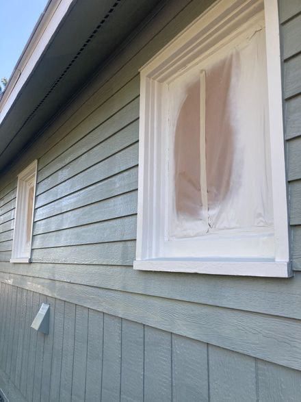 Two windows on a blue-gray shingled house wall are covered with white protective plastic for painting.