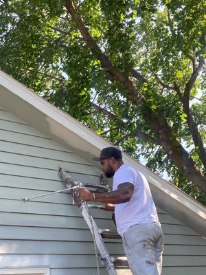 A person on a ladder uses a paint sprayer to apply light-colored paint to the exterior siding of a house.