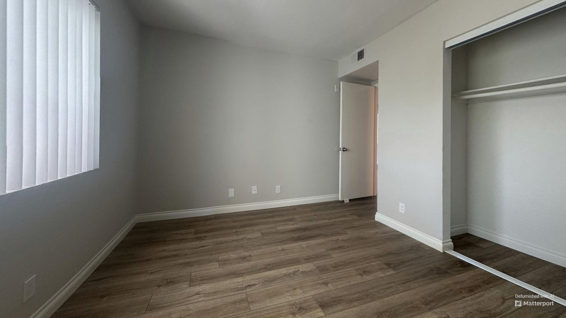 Empty bedroom with wooden floor, closet, closed door, and window with blinds.