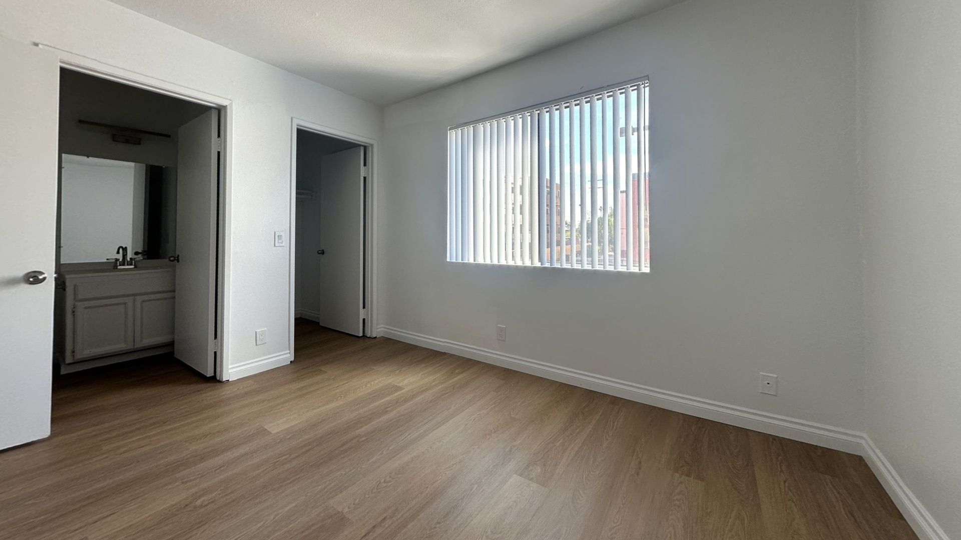 Empty bedroom with wood-look flooring, white walls, and a window with blinds. Doors lead to a bathroom and another room.