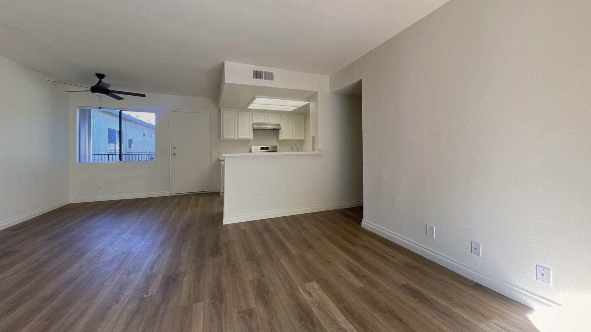 Empty apartment interior with wood-look flooring, white walls, and a partially visible kitchen area.