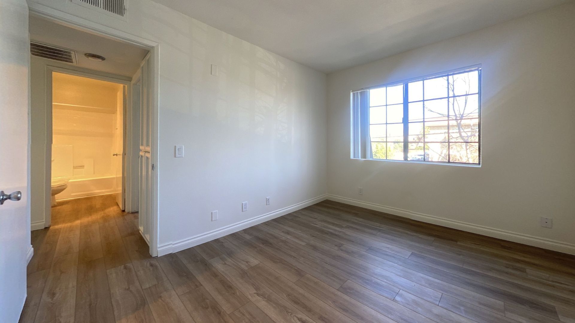 Empty bedroom with wood-look flooring and a window. Doorway leads to a bathroom.