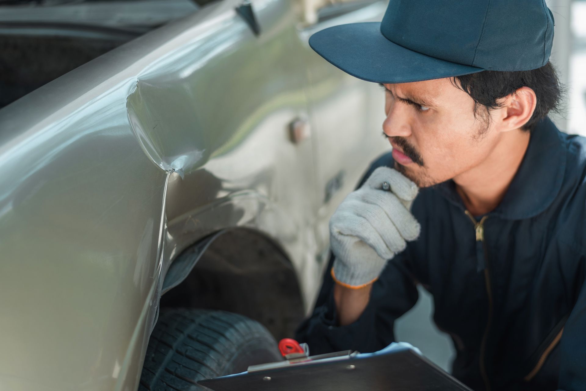 A man is looking under the fender of a car while holding a clipboard.