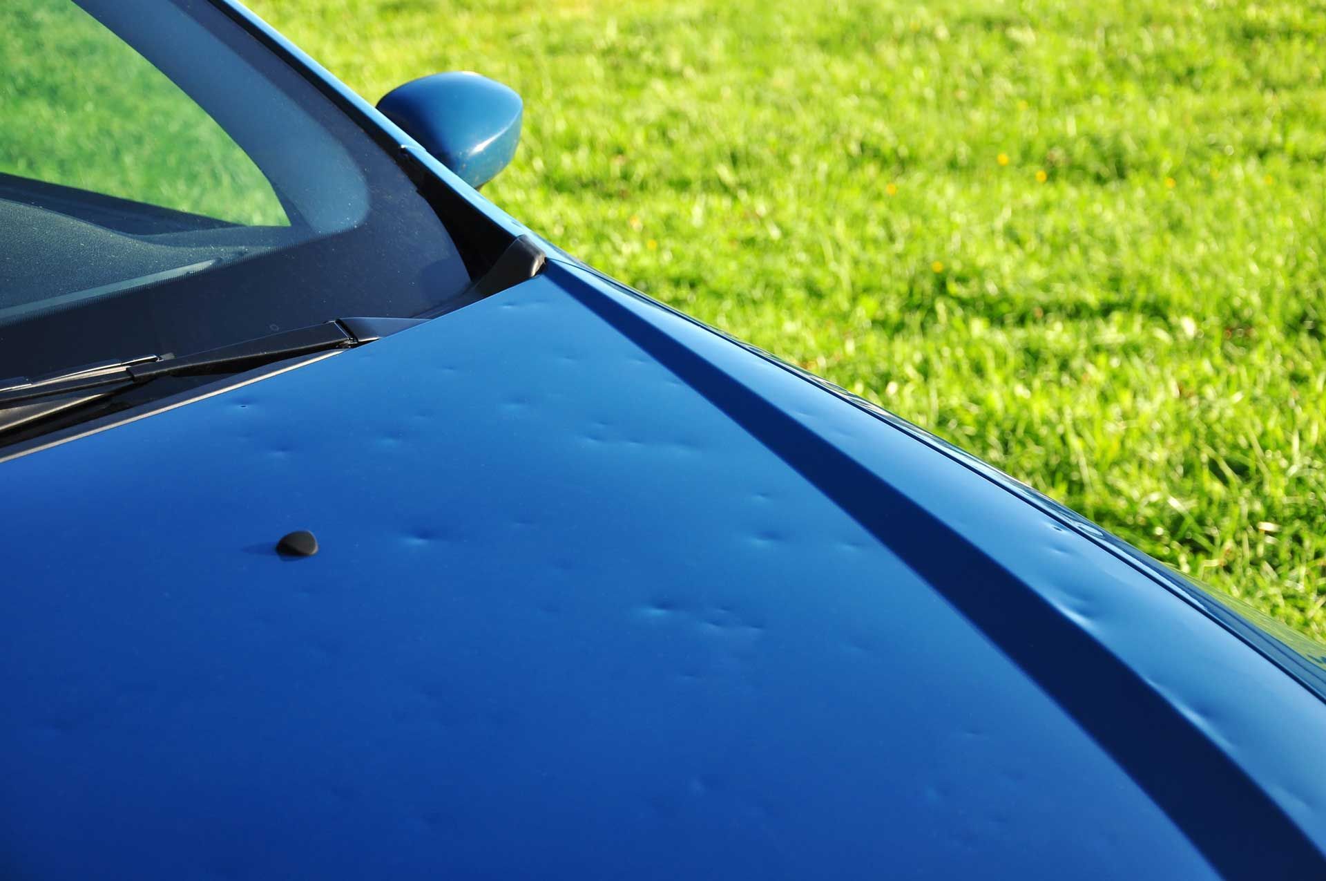 A blue car with a damaged hood is parked in a grassy field