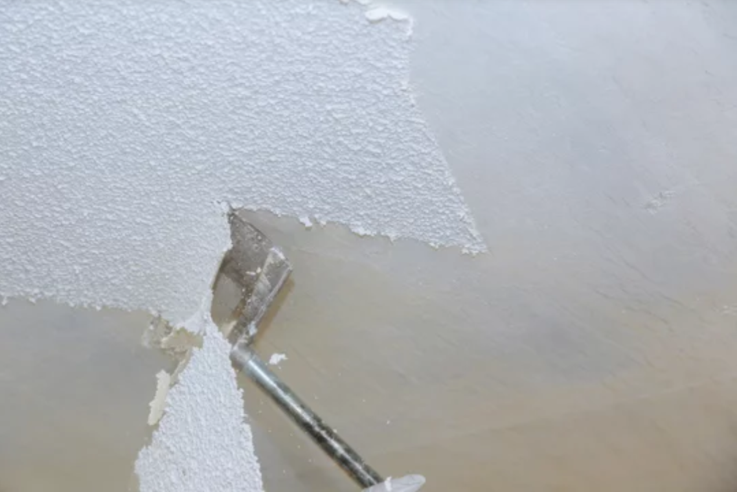 Worker removing old, dirty popcorn ceiling texture from a wall with a scraper tool.