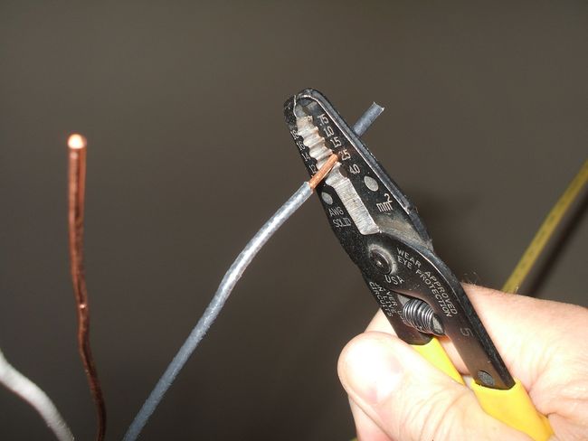 Hand stripping insulation from a gray wire with pliers beside a copper wire end