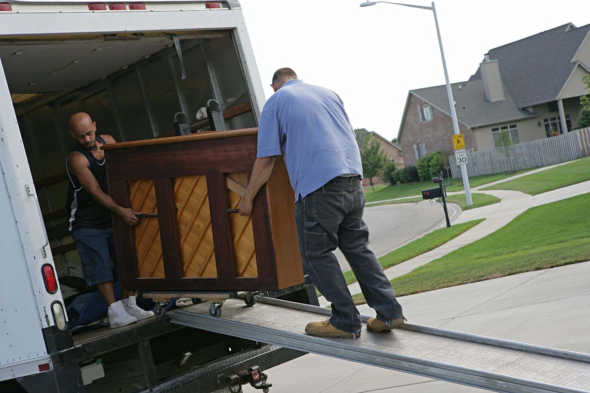 Two men are loading a wooden upright piano onto a truck during a piano moving job with a ramp.