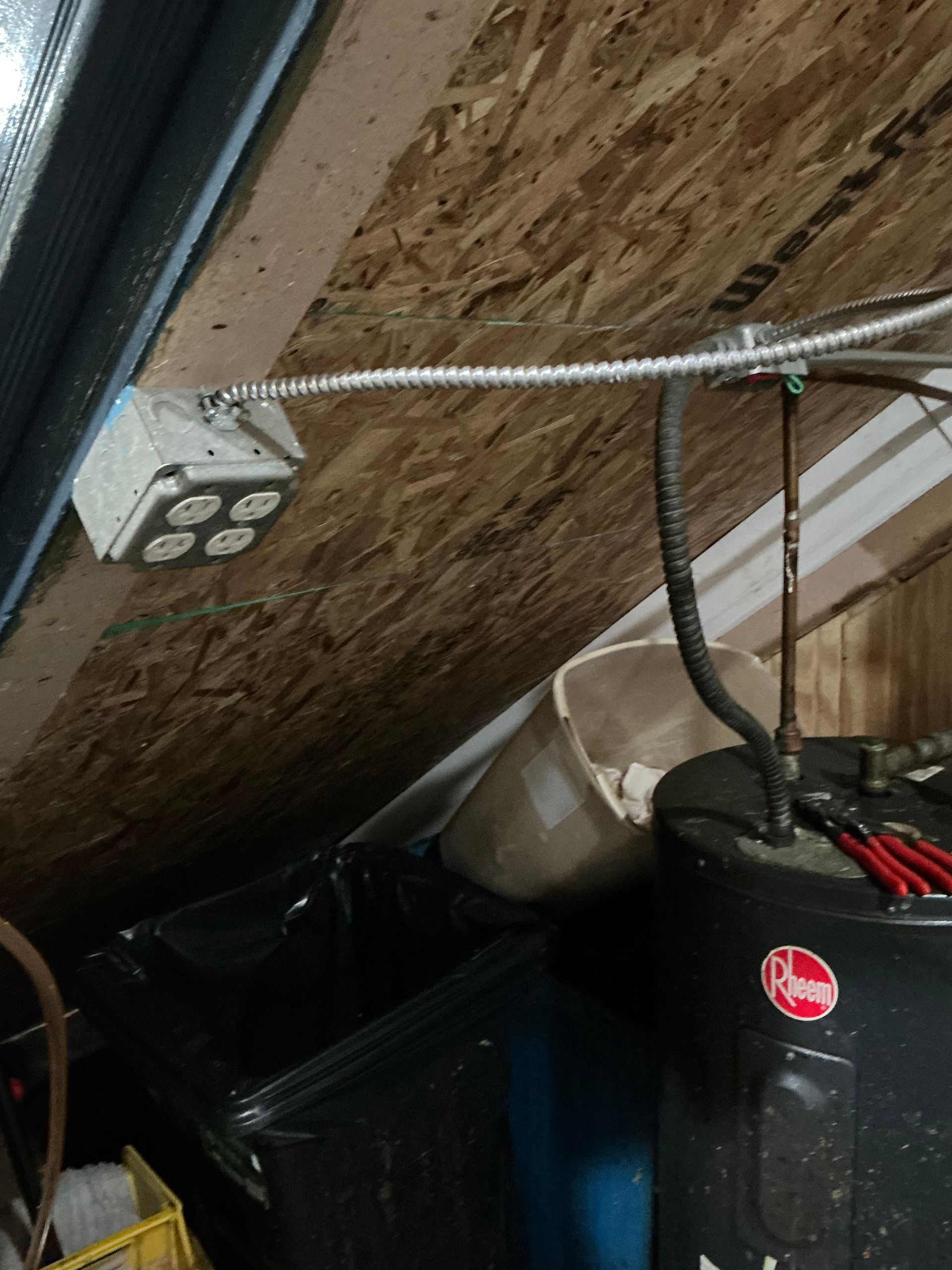 Electrical outlet box mounted on angled wooden paneling, near a water heater and storage.