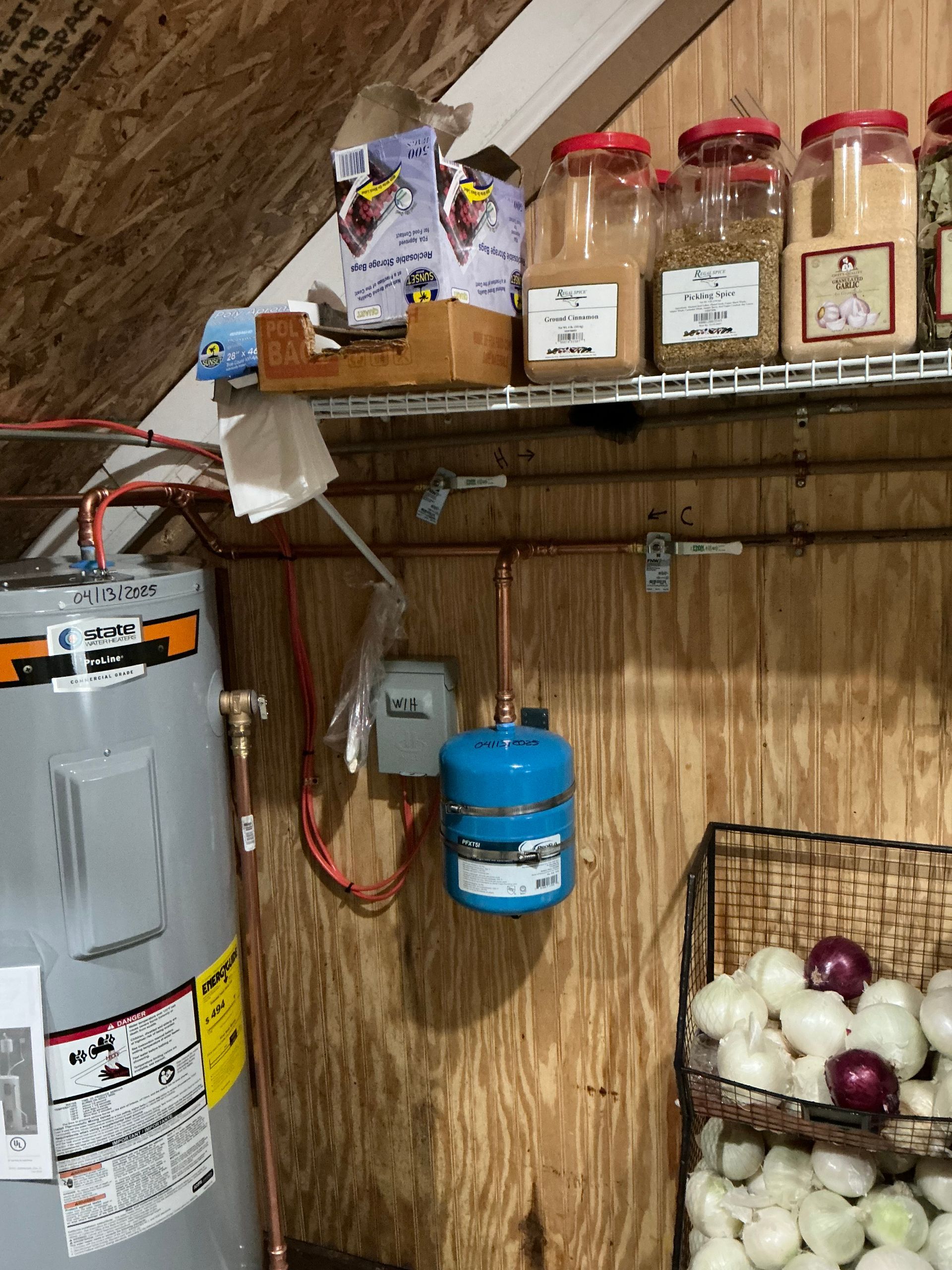 Water heater, expansion tank, shelves with spices, onions in basket, against wood paneling.