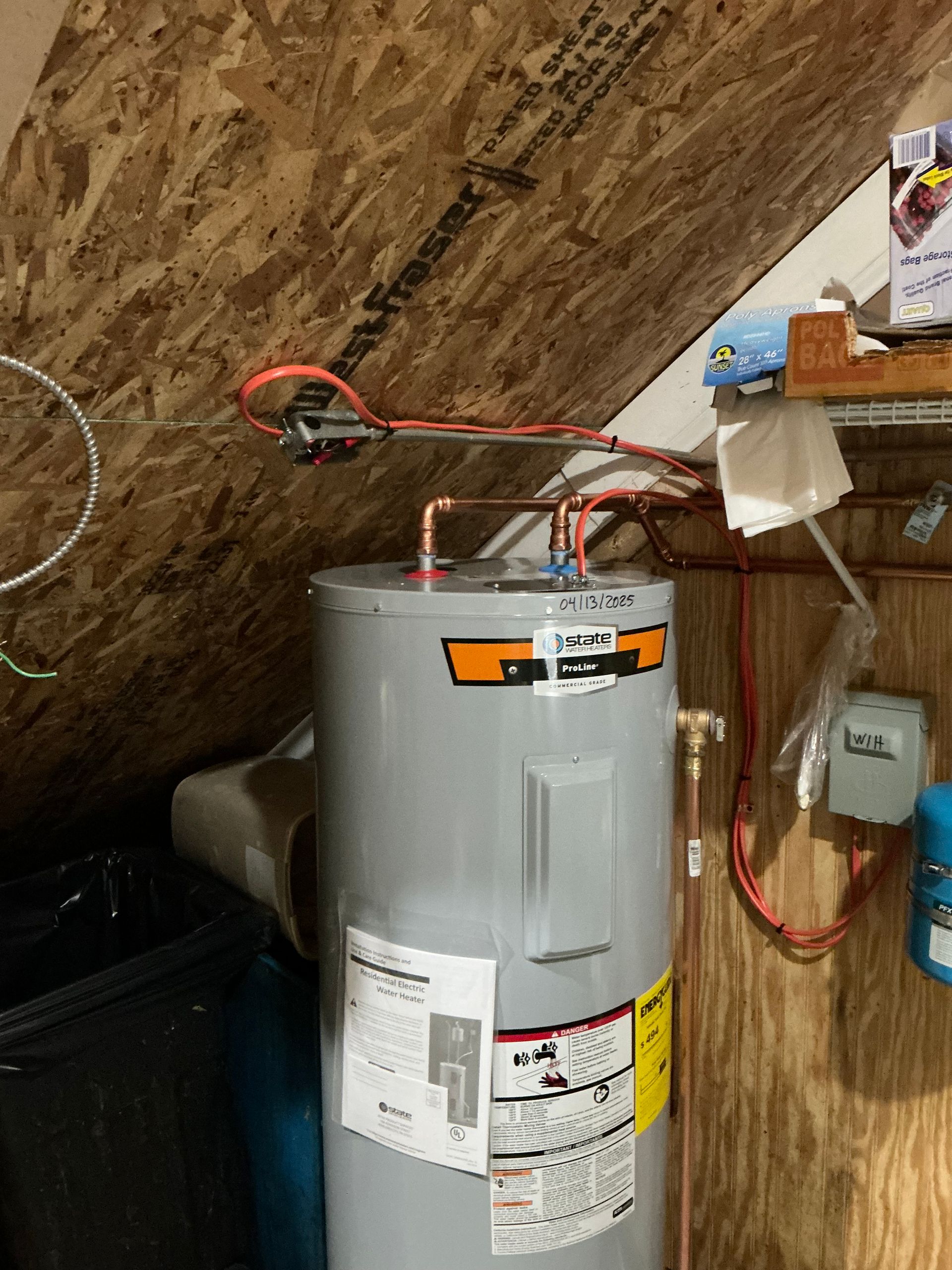 A tall gray water heater in a wooden room, with copper pipes connected and warning labels visible.