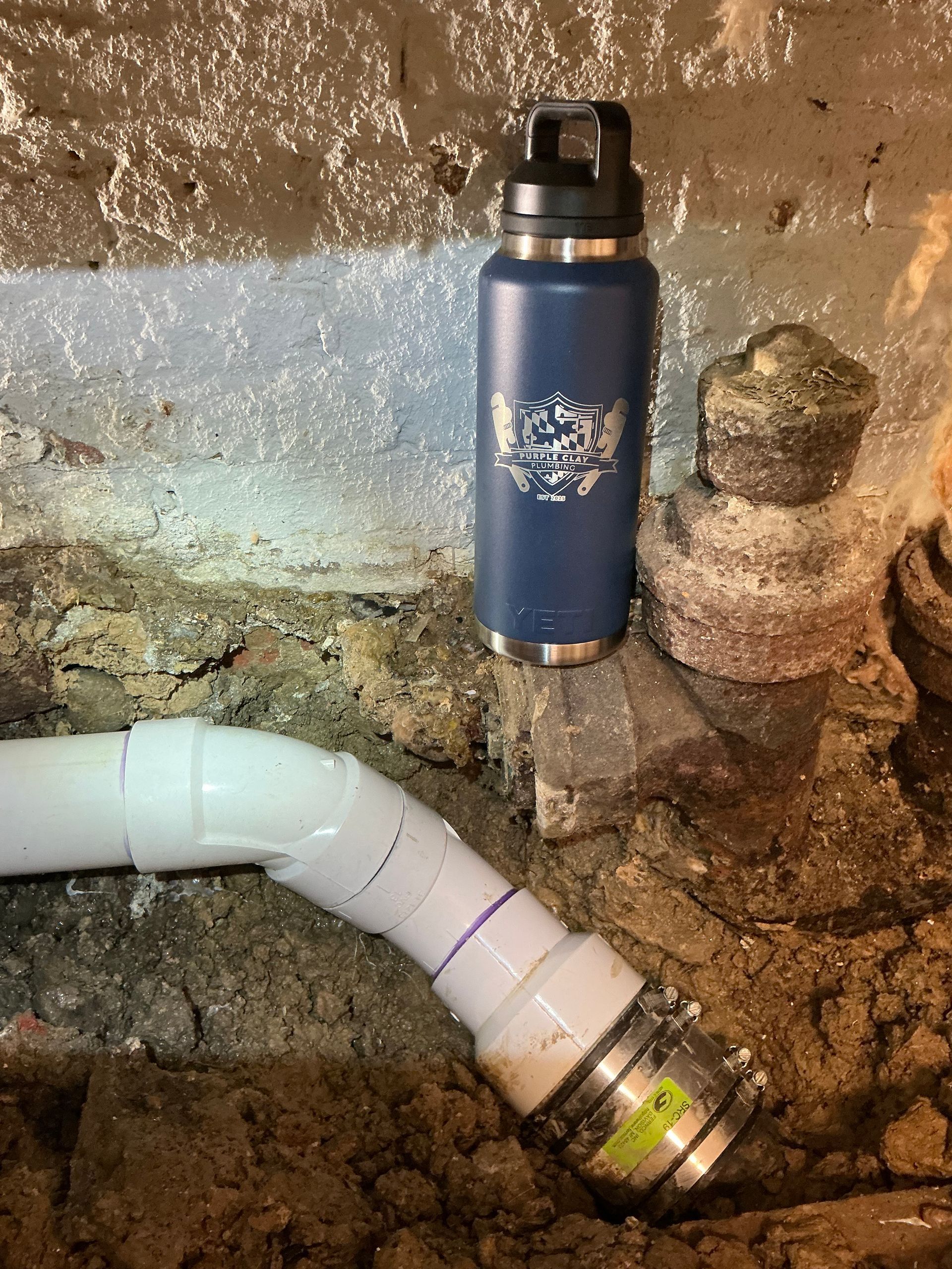 Blue water bottle next to white PVC pipe with a metal fitting in a dirt basement.