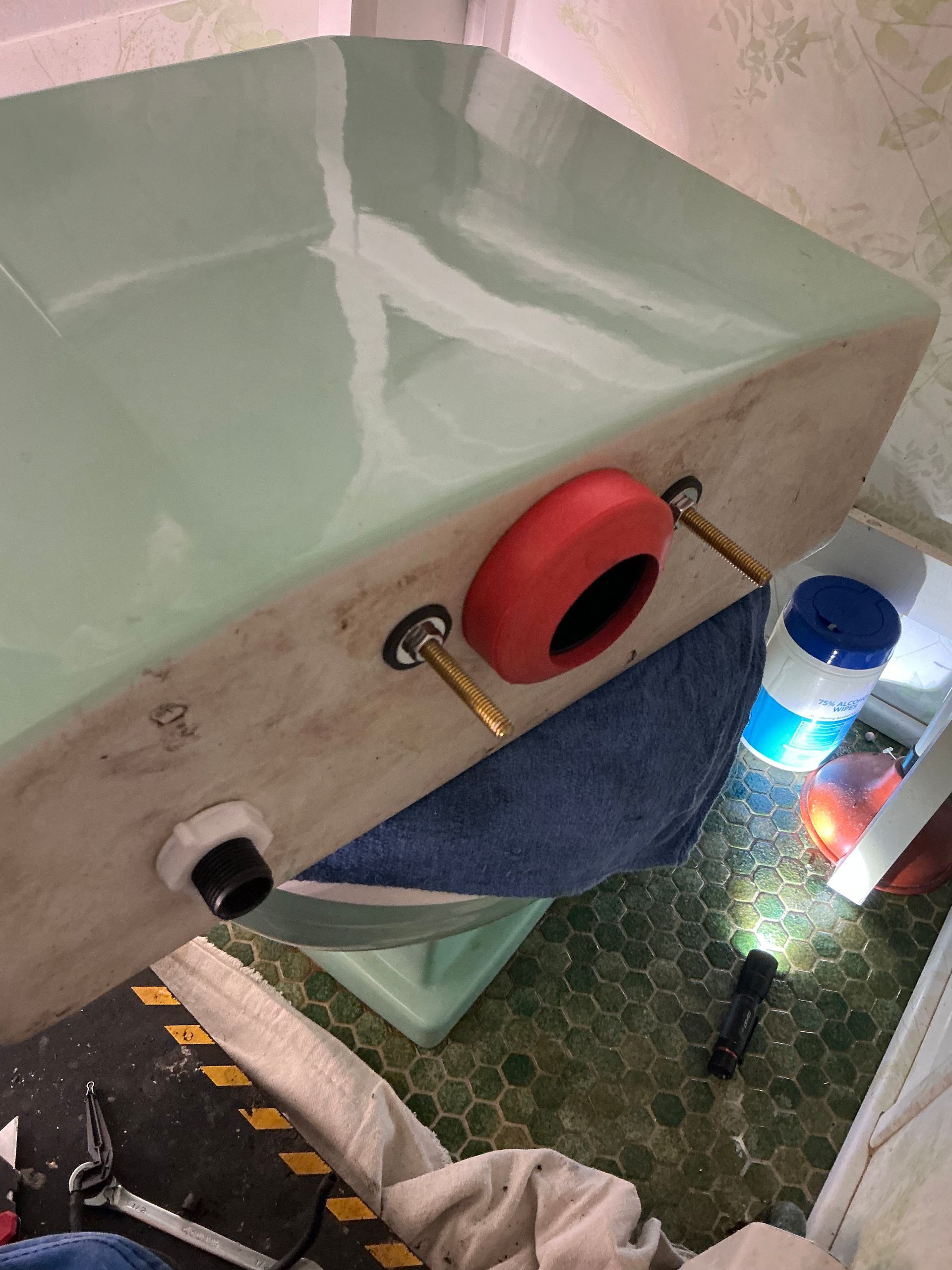 Light green toilet tank with a red flapper, bolts, and a black flush valve. On a green tiled floor.