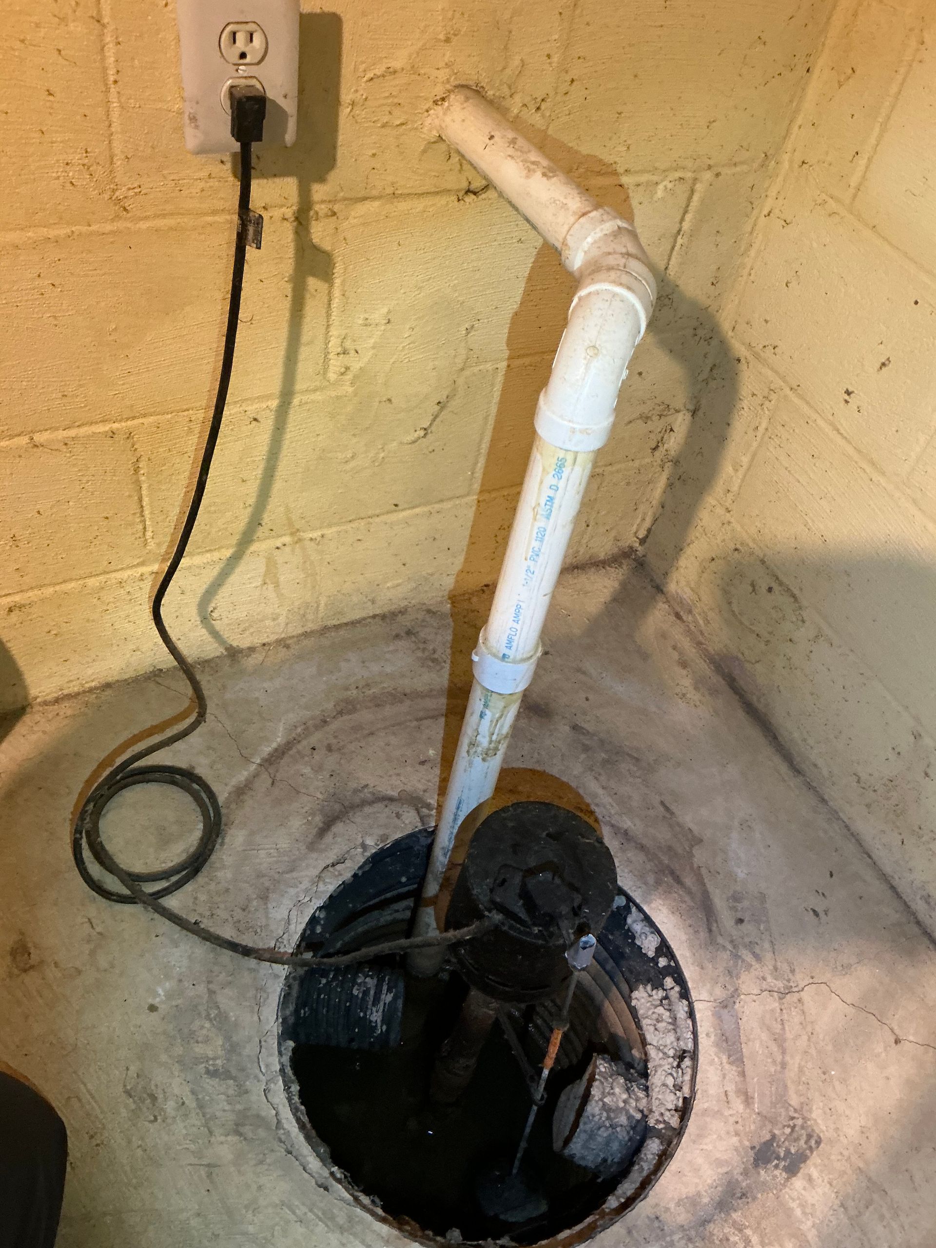 Sump pump in a circular pit with a white PVC pipe, plugged into an outlet on a concrete wall.