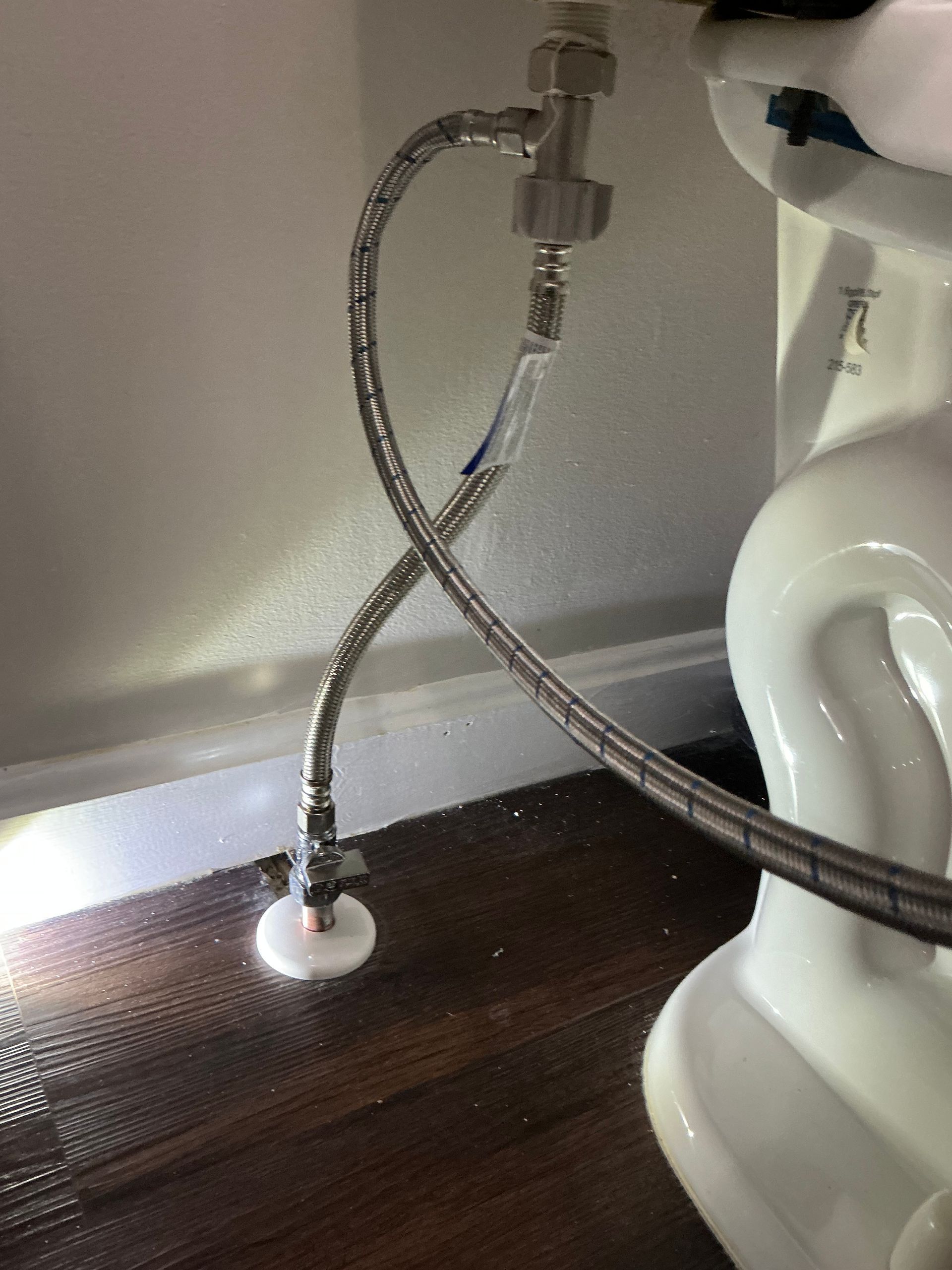 Toilet with plumbing visible: silver braided hoses crossing over wood flooring near a white wall.