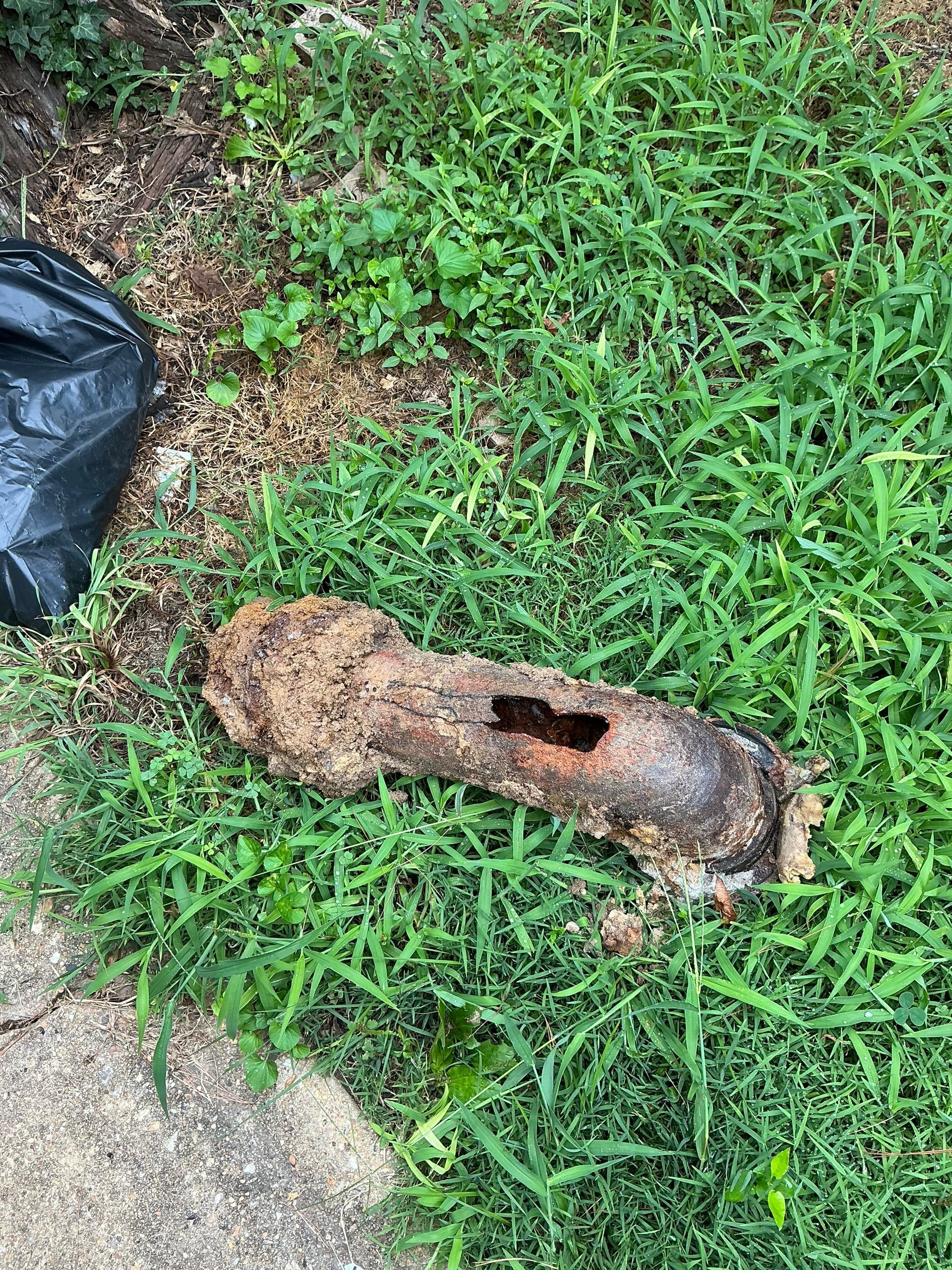 Brown, damaged pipe on grass next to a sidewalk and black trash bag.