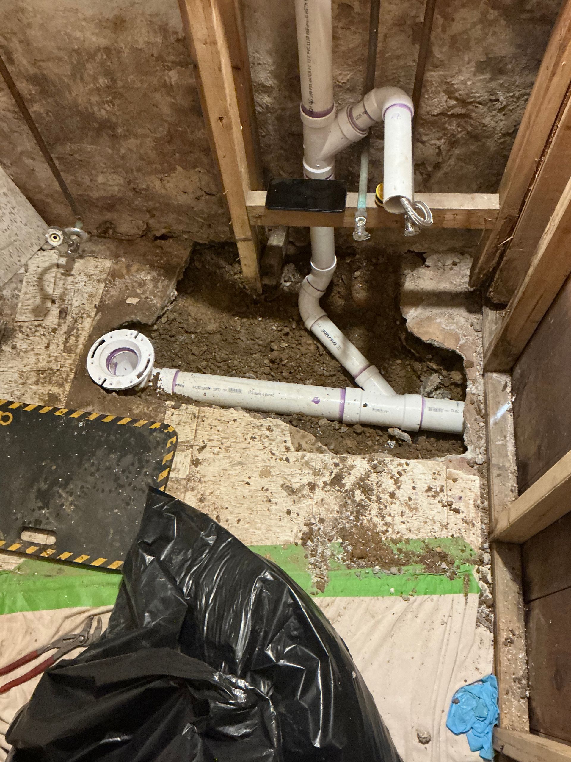 Overhead view of exposed plumbing, pipes, and a drain in a construction setting. White and purple PVC pipes are visible.