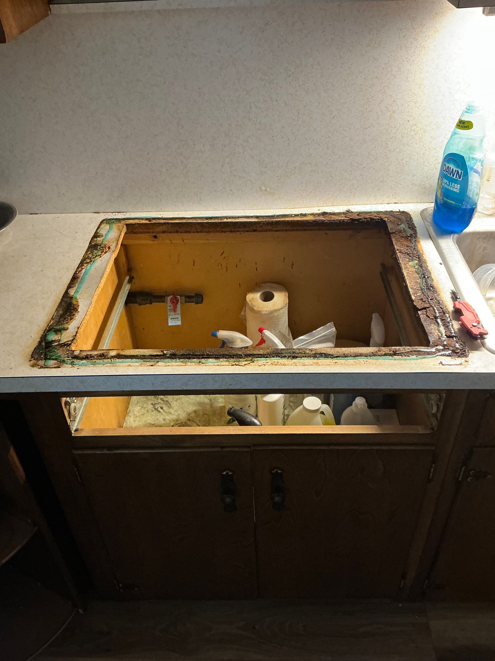 Open cabinet under a countertop with a removed sink; cleaning supplies and clutter visible.