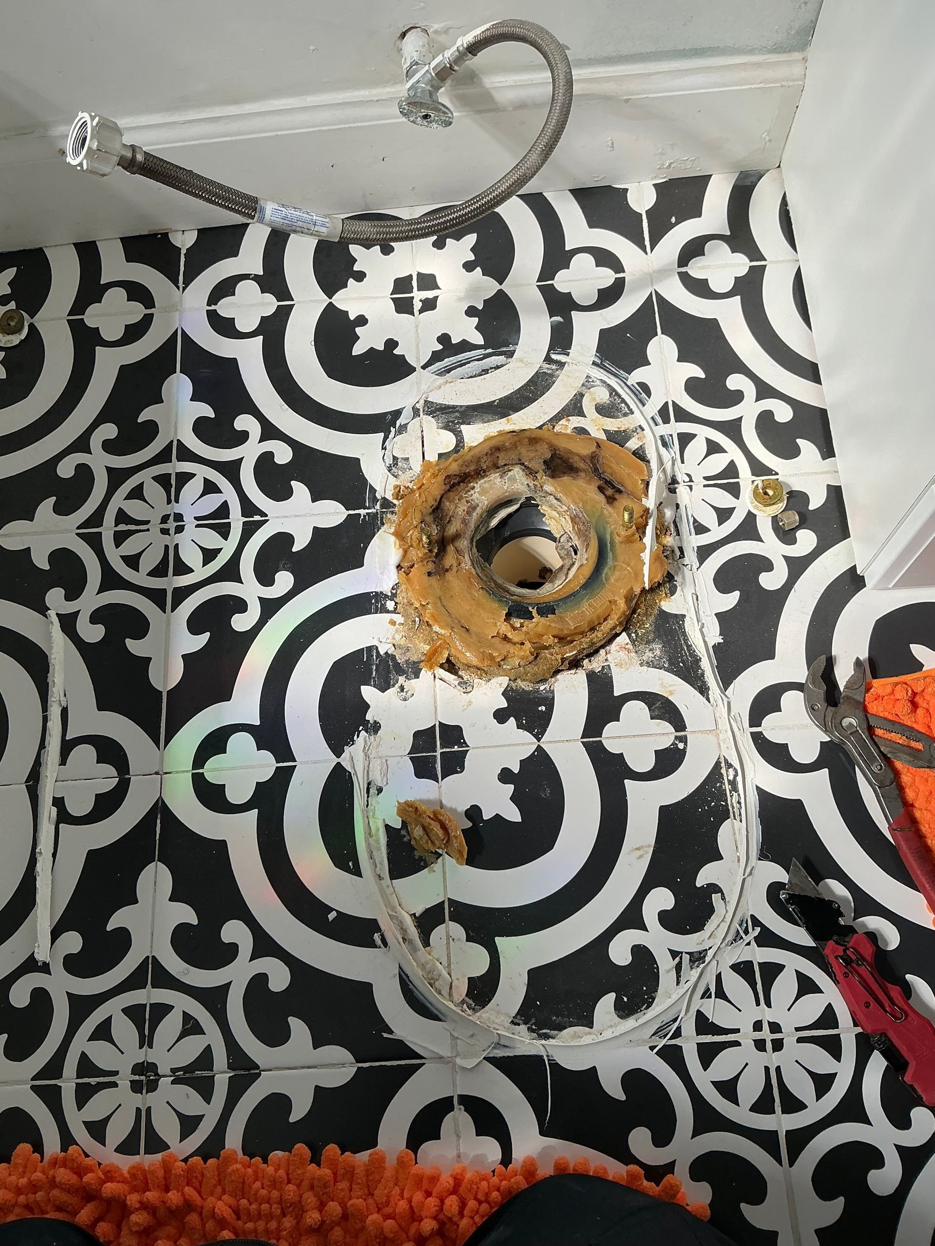 Toilet flange repair on patterned black and white tiled floor. Brown wax ring and plumbing exposed.