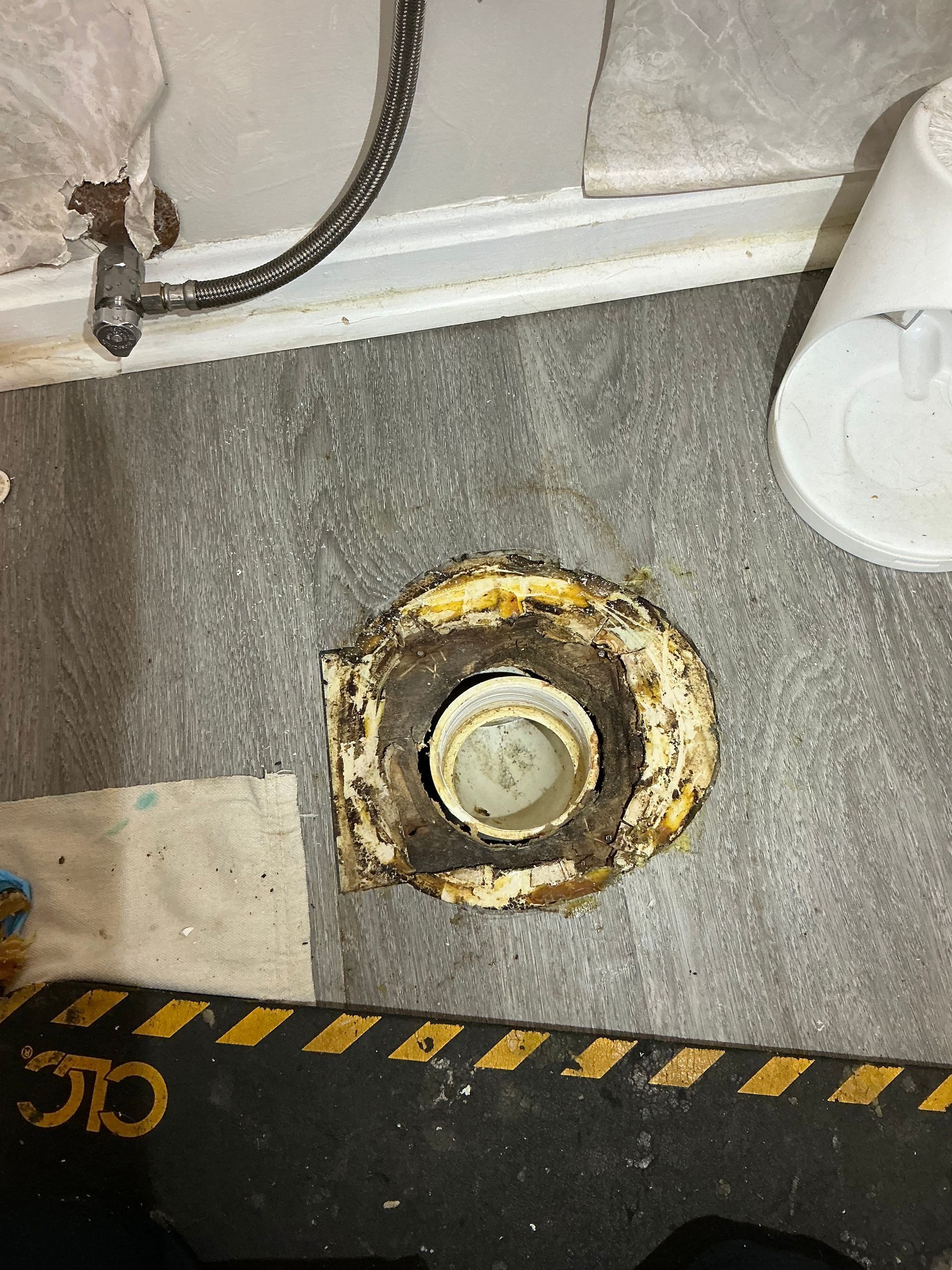 Floor drain surrounded by debris and a partially visible black and yellow caution sign.