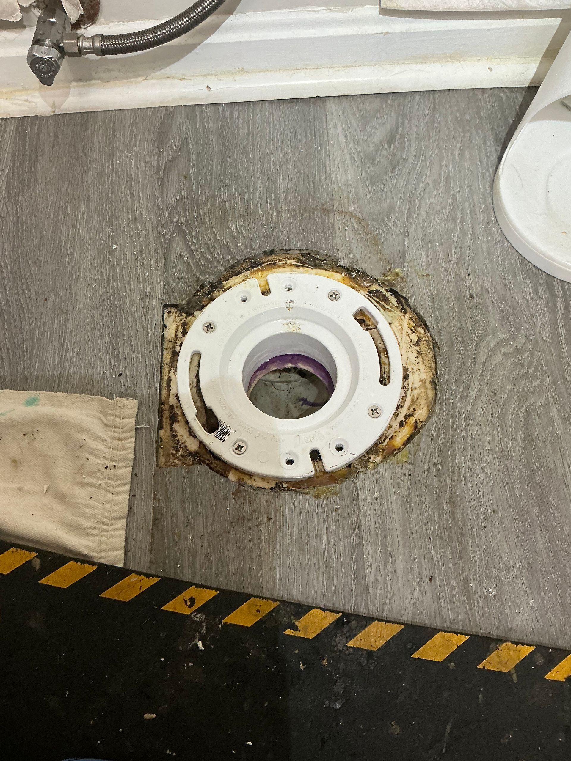 Toilet flange installed on gray vinyl flooring. Black and yellow hazard tape visible.