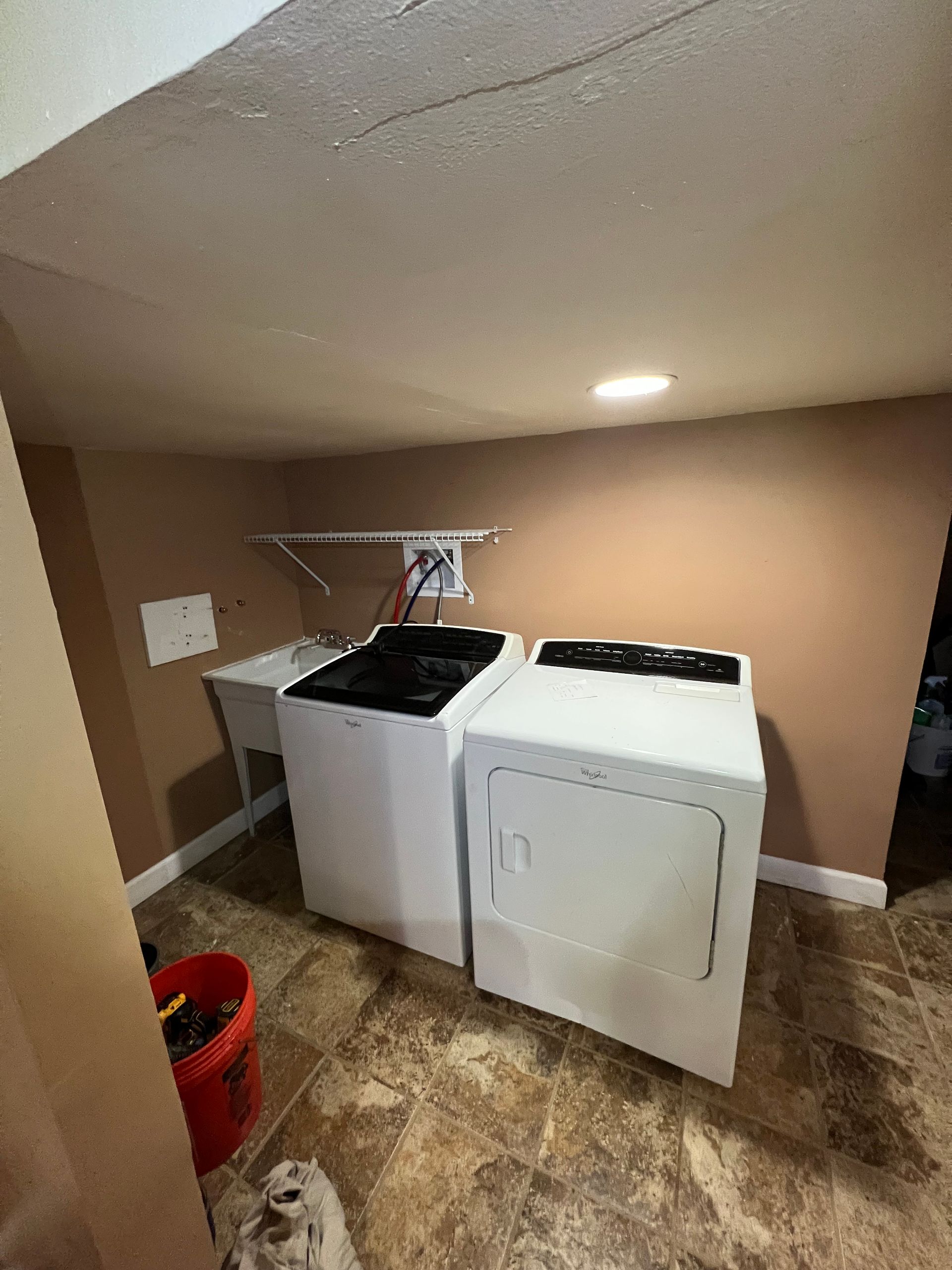 Laundry room: white washer and dryer, tan walls, utility sink, and red bucket.