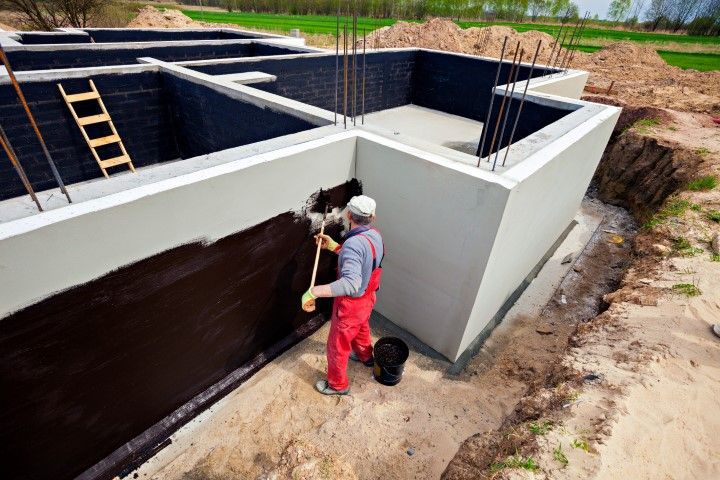someone on a construction site applying black sealer to the exterior of a basement foundation