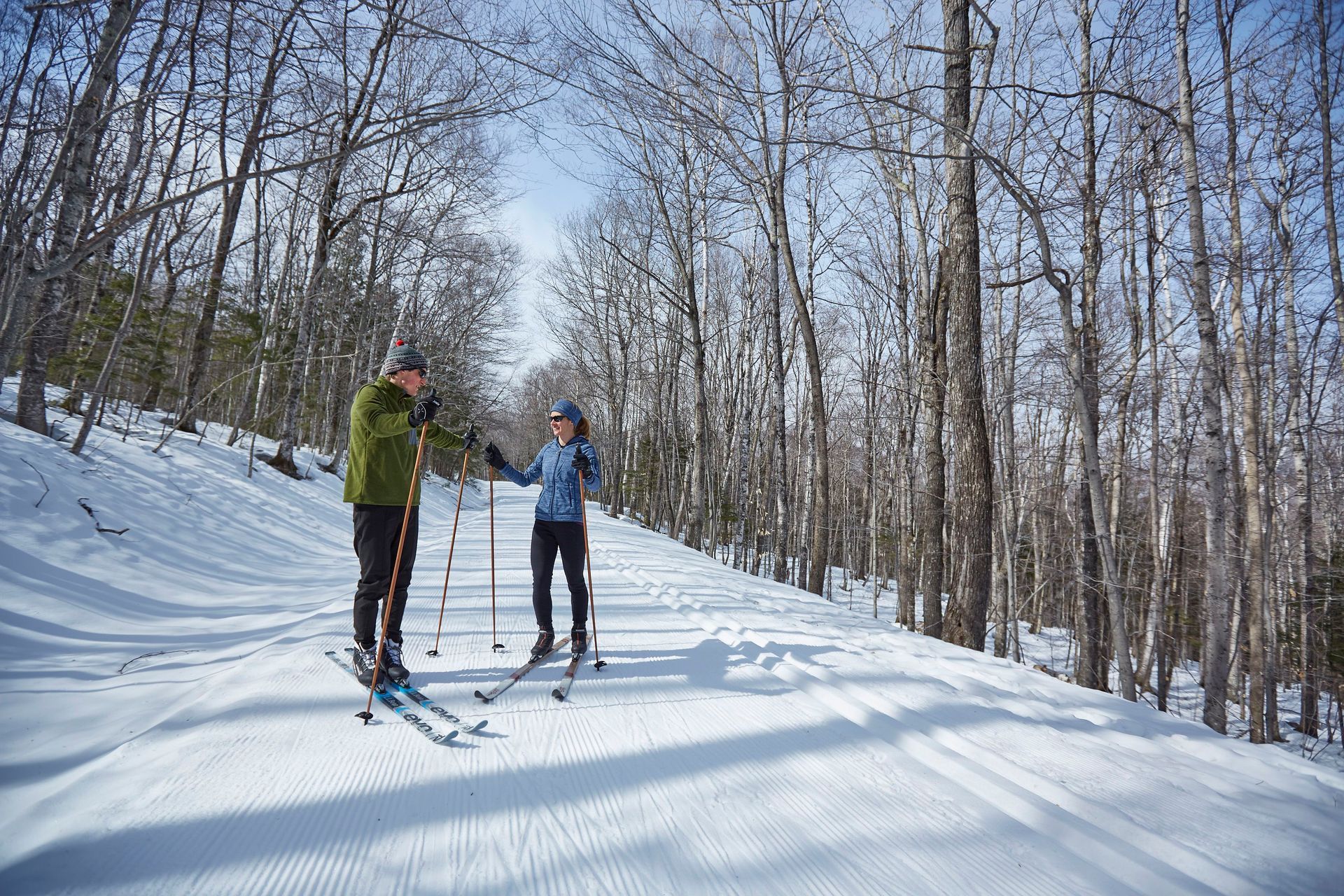 A man and a woman on cross-country skis, stopped on a groomed trail, talking.