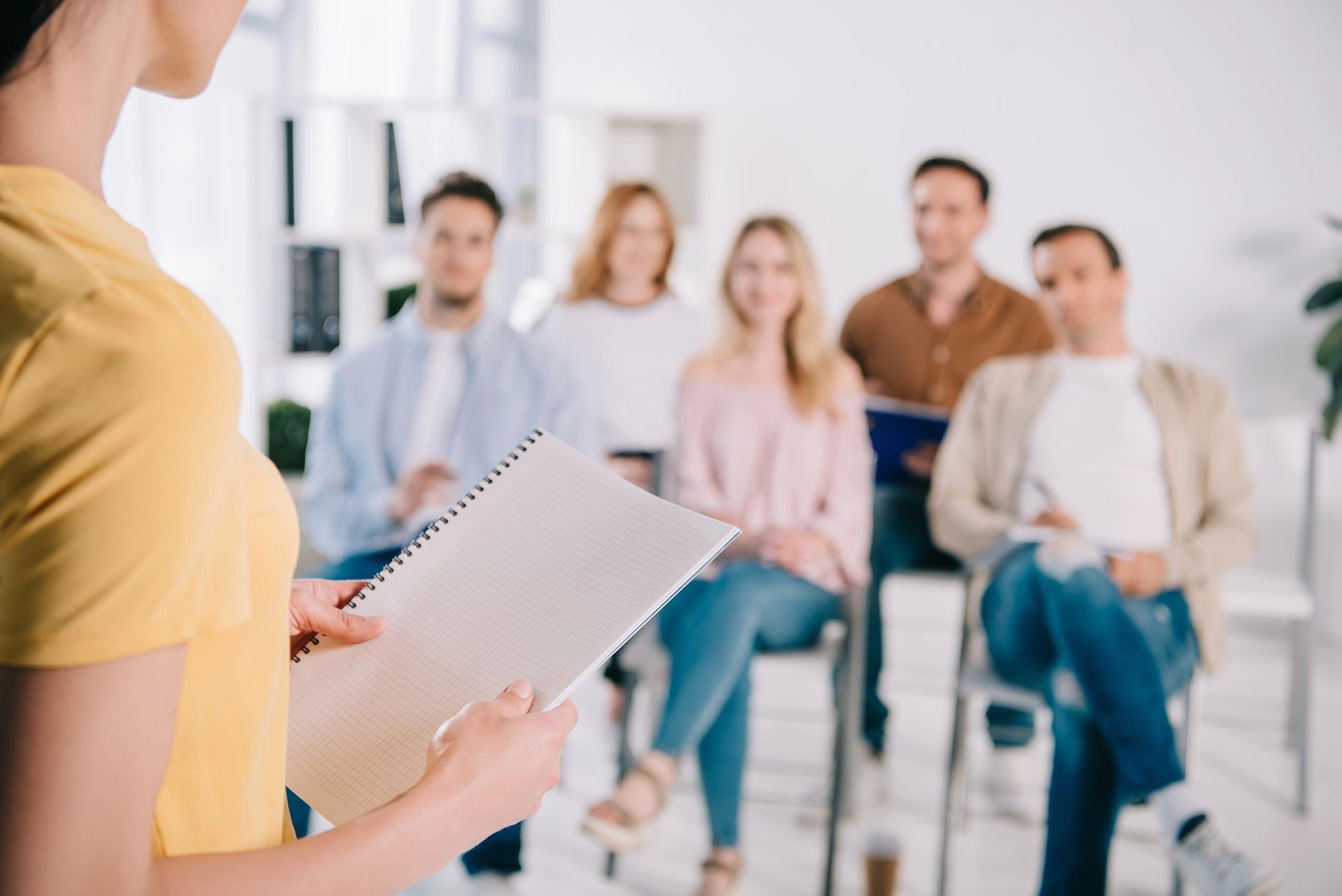Mujer haciendo una presentación ante un grupo sentado en una habitación luminosa; sostiene un cuaderno.