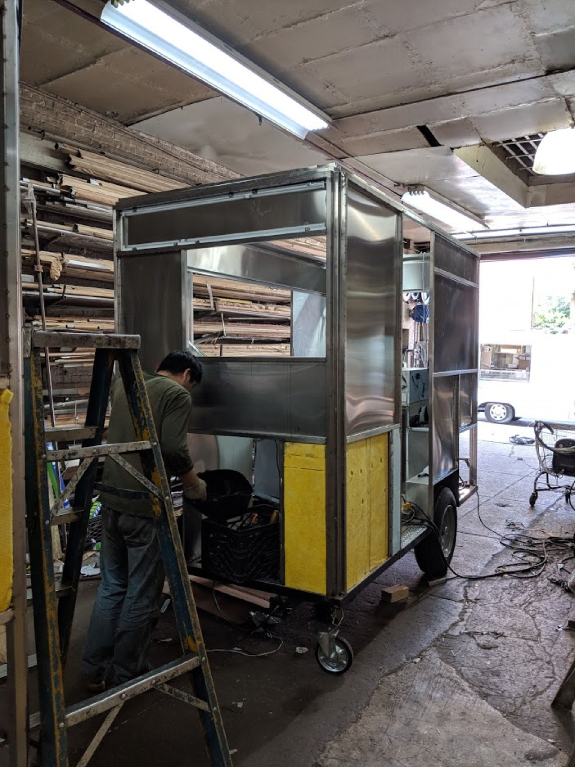 A man is working on a stainless steel food truck in a garage.