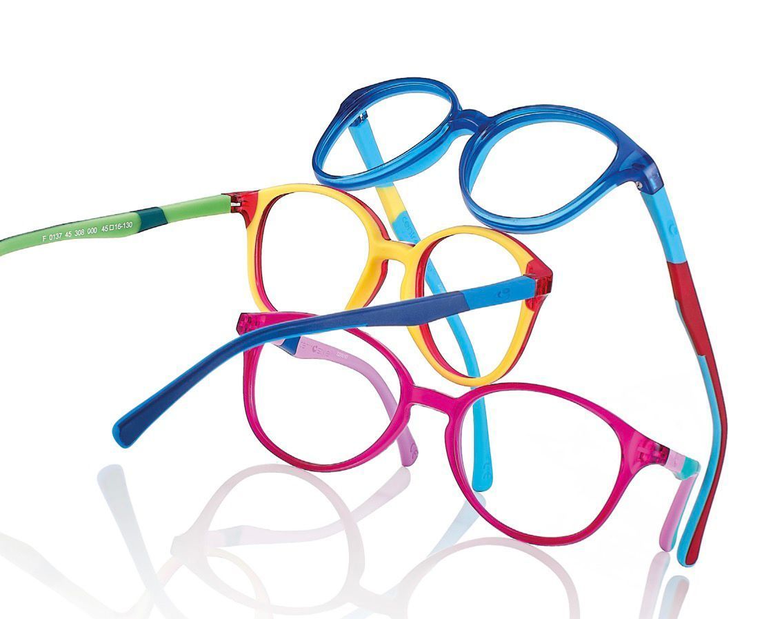 Un paio di occhiali geometrici esposti su uno scaffale rosa in un negozio. Montatura nera con lenti trasparenti e un'immagine promozionale sullo sfondo.