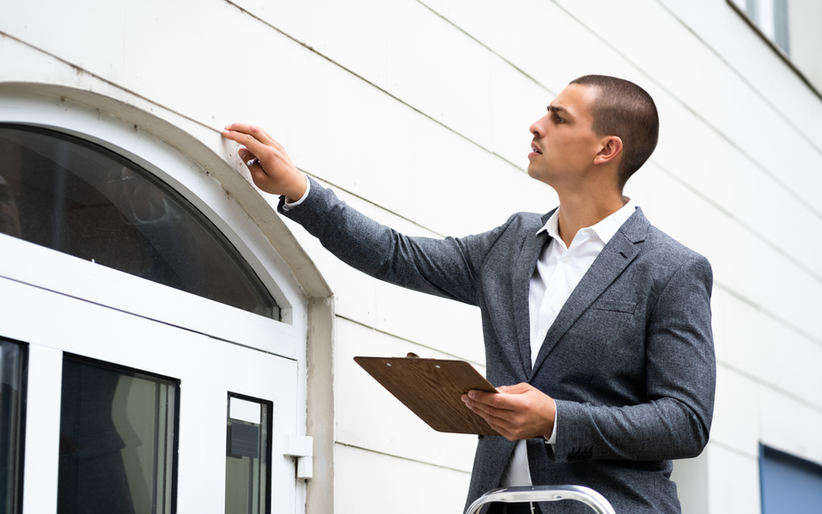 Man in a gray blazer inspecting the exterior of a white building and holding a clipboard