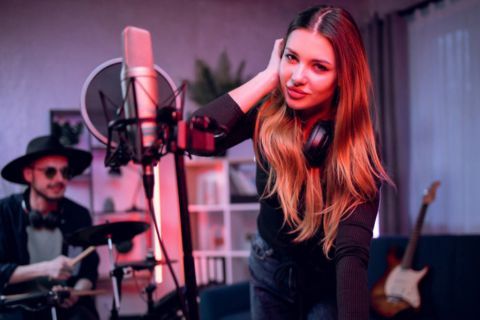 A woman is singing into a microphone in a recording studio while a man plays drums.