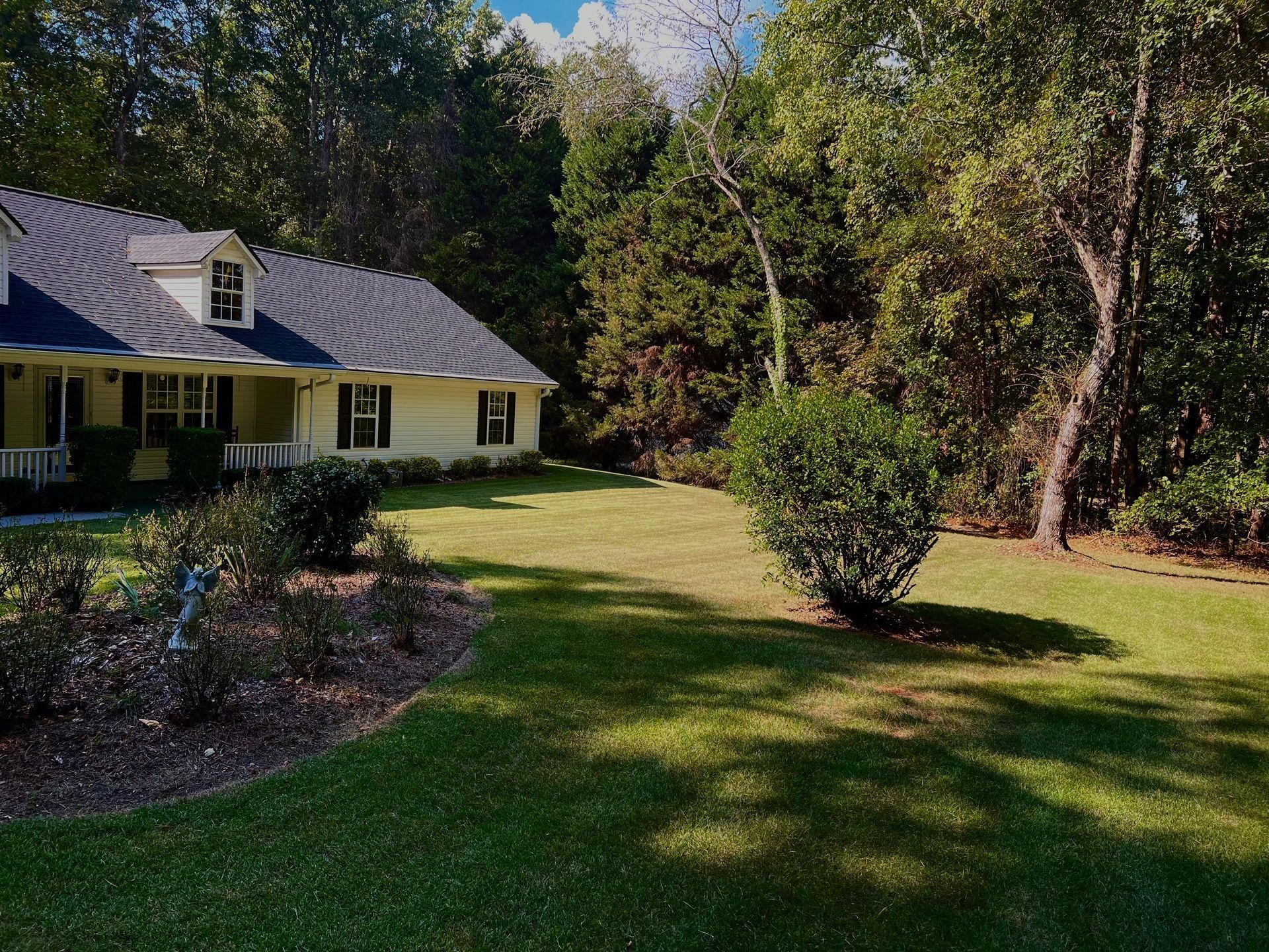 Yellow house with dark roof beside a green lawn and trees in a sunny yard