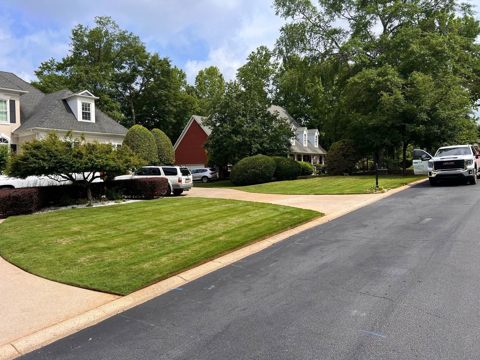 Quiet suburban street with houses, green lawns, trees, and parked cars on a sunny day
