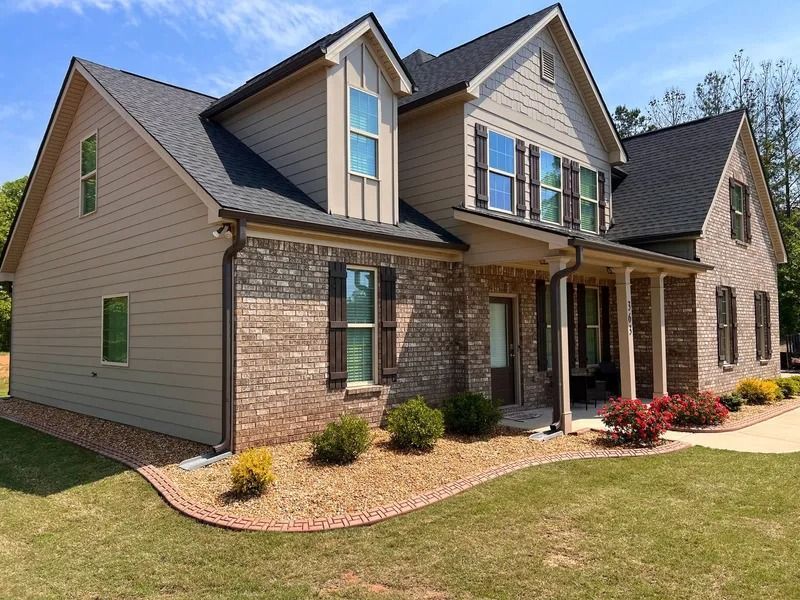 Two-story suburban brick house with beige siding, gray roof, and front porch in a landscaped yard