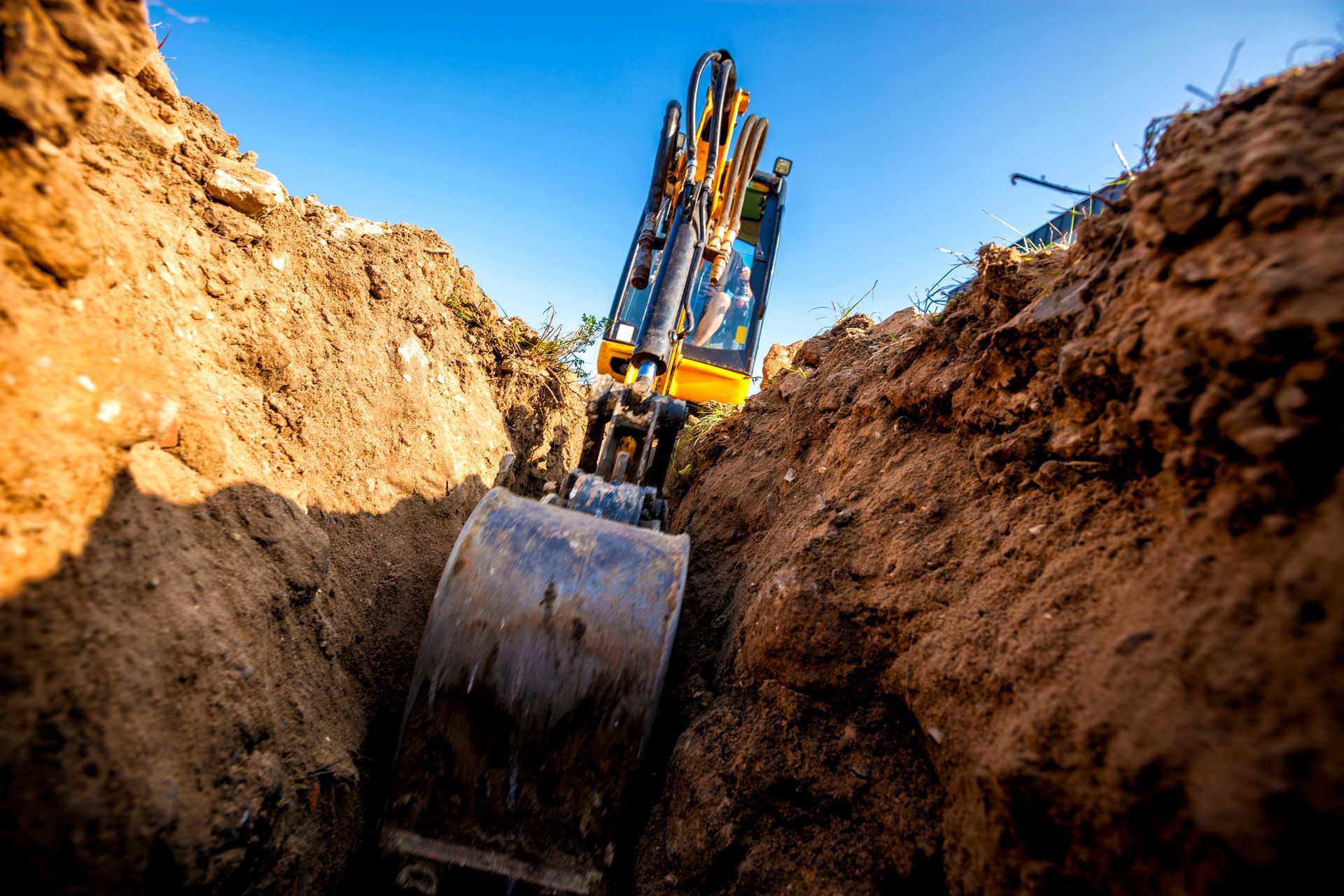 Excavator digging a trench in brown earth against a clear blue sky.