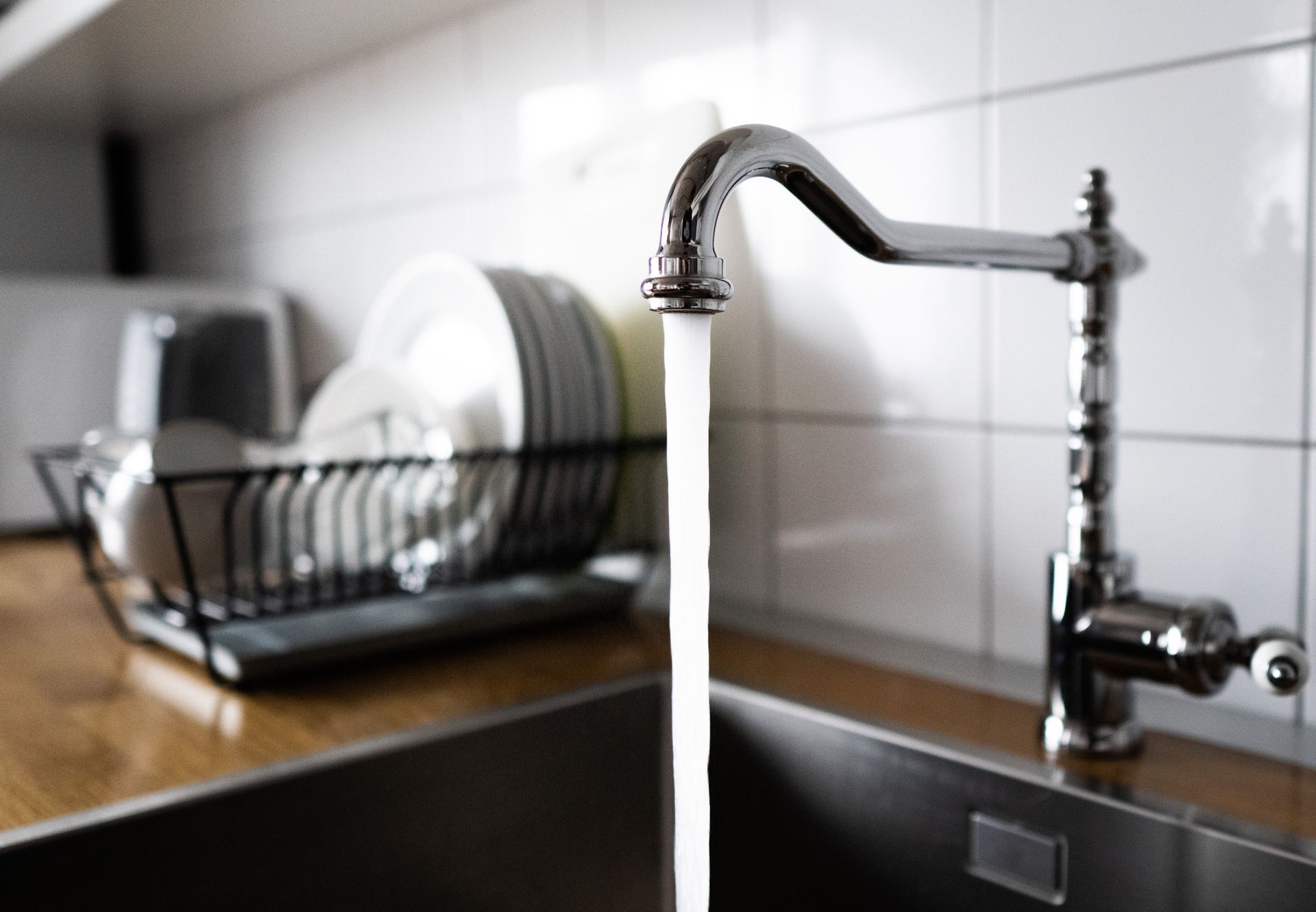 Faucet with water running into a stainless steel sink, near a dish rack and white tiled wall.