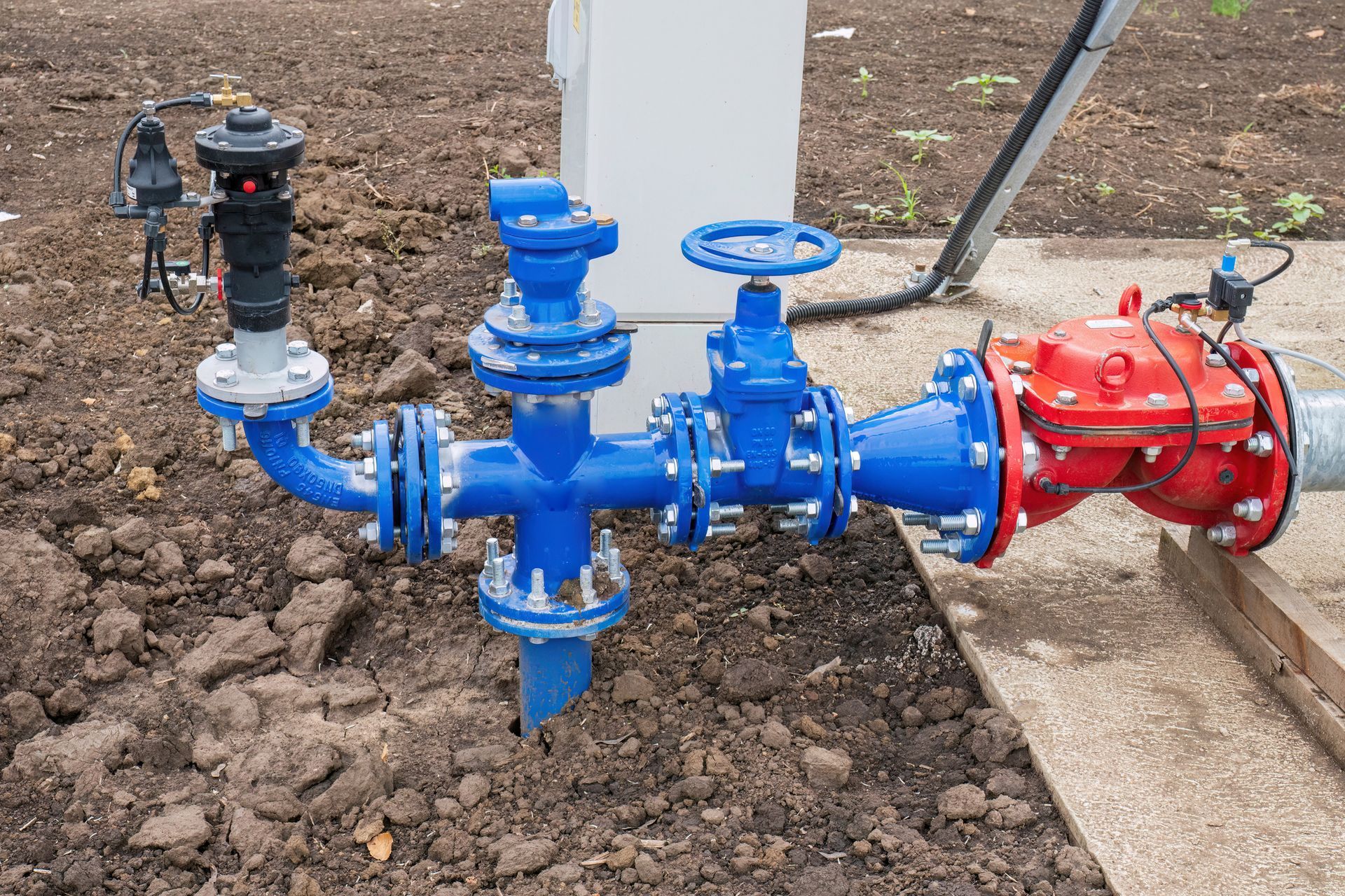 Blue and red industrial water pipes and valves, set in dirt, connected to a white post.