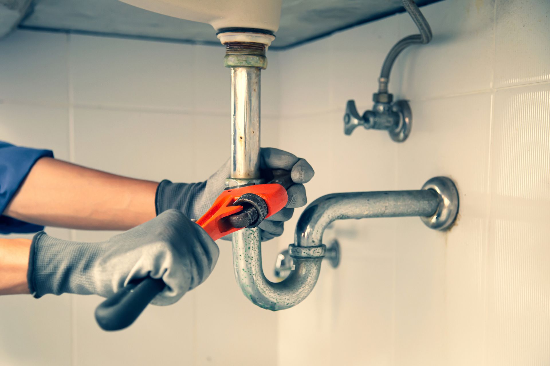 Plumber in gray gloves using a wrench to work on a curved pipe under a white sink.