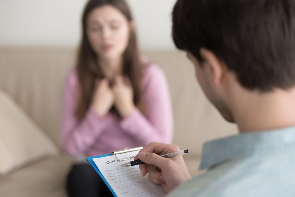 Therapist taking notes during a session with a woman, who is sitting on a sofa with eyes closed and hands on chest.