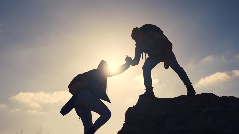 Hikers helping each other climb a rocky summit, silhouetted against a bright sky.