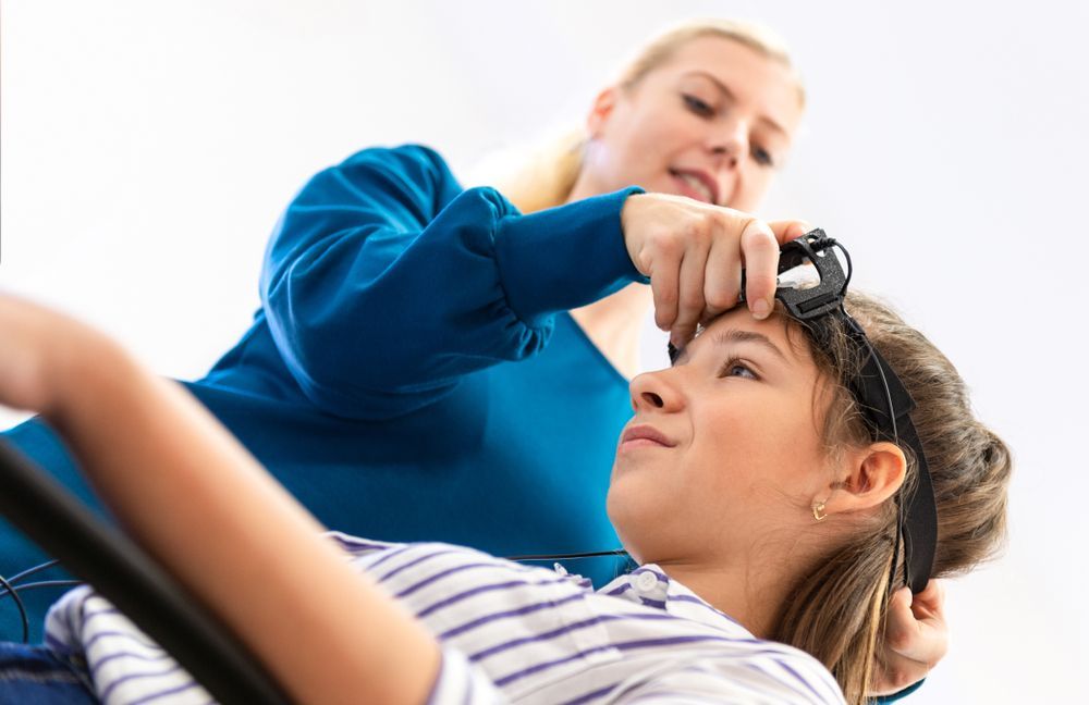 Woman adjusting a headband on a girl's head; both are indoors, the girl is lying down.