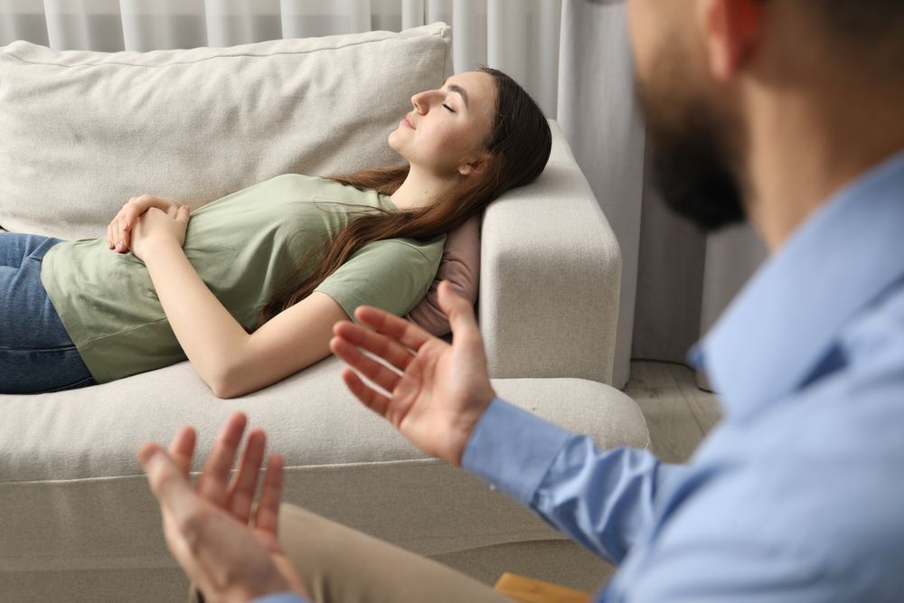 Woman lying on a couch, eyes closed, during a therapy session, with therapist gesturing.