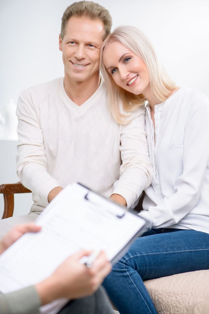 Couple smiling, sitting together, looking at a person holding a clipboard and pen.
